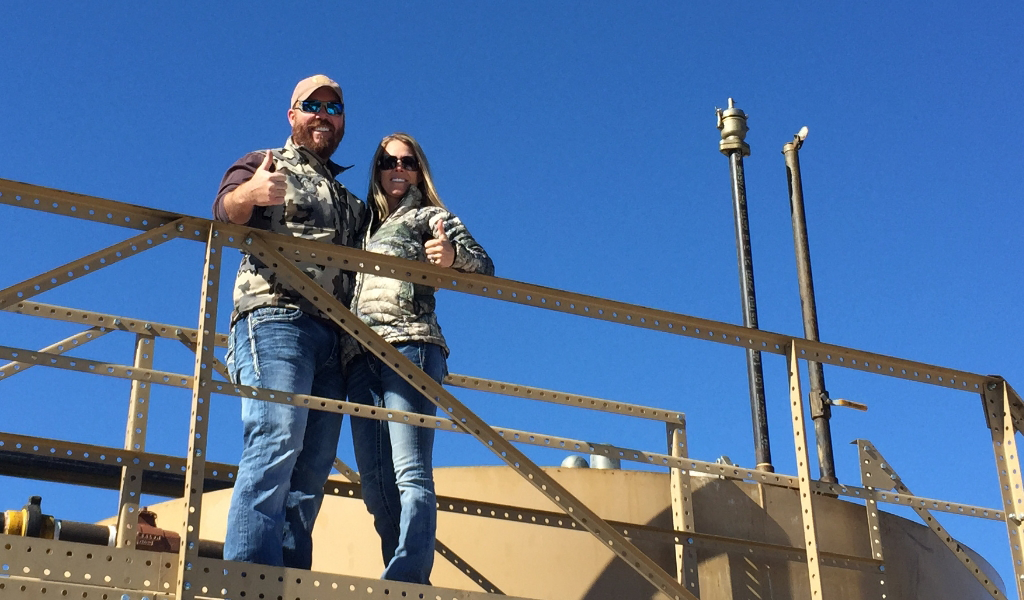 Two people in camouflage jackets stand on a metal platform, giving thumbs up, against a clear blue sky.
