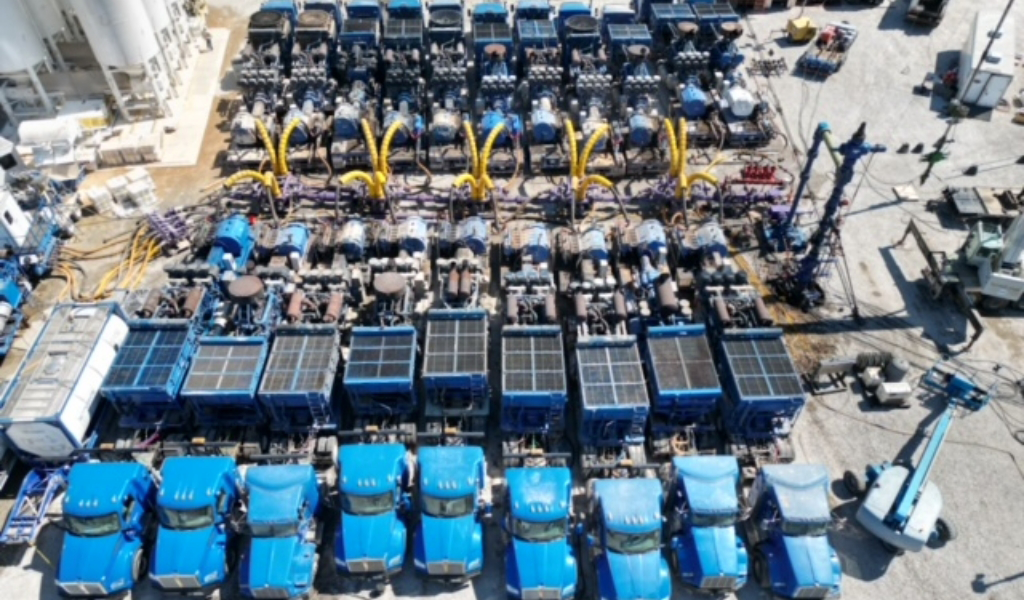 Aerial view of multiple blue trucks and connected machinery arranged in rows at an industrial site, with yellow hoses extending from the equipment.