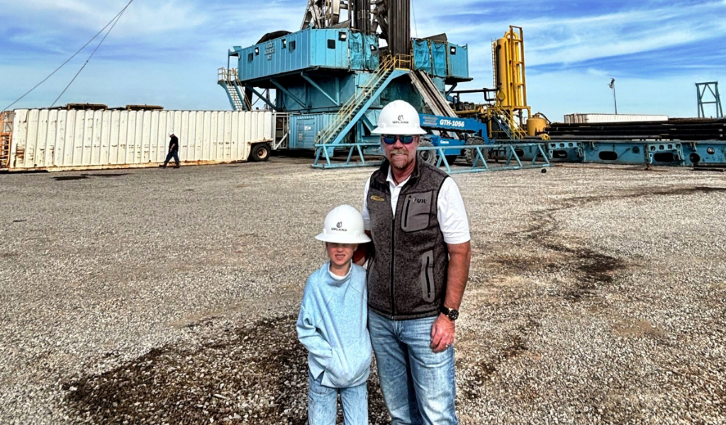 An adult and a child wearing hard hats stand on a gravel surface in front of industrial machinery.