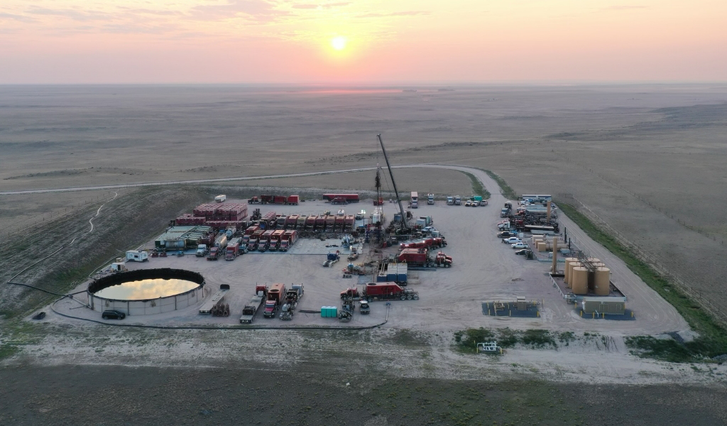 Aerial view of an oil drilling site at sunset with equipment, storage tanks, and a circular containment area, set against a vast plains landscape.