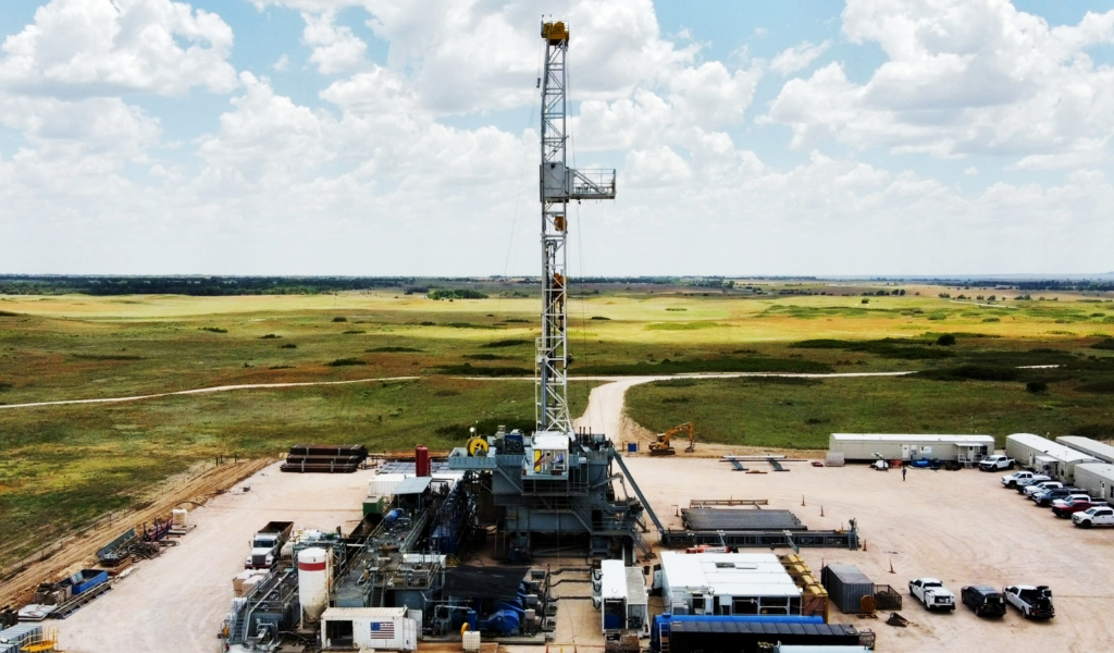 A large oil drilling rig is situated in an open field under a cloudy sky, surrounded by equipment, vehicles, and containers.
