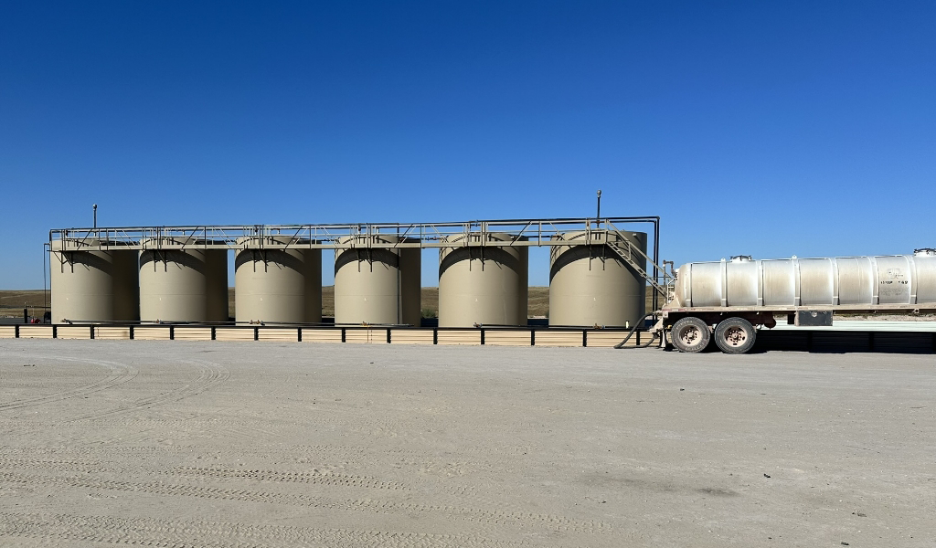 A row of six large cylindrical storage tanks is lined up beside a parked white tanker truck on a clear day under a blue sky.