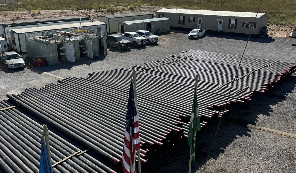 Stacks of metal pipes arranged in rows on gravel, with several vehicles parked nearby. Prefabricated buildings and flags are also visible in the background.