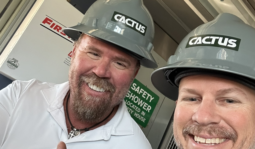 Two men wearing gray hard hats labeled "CACTUS," smiling, stand indoors in front of a safety sign.