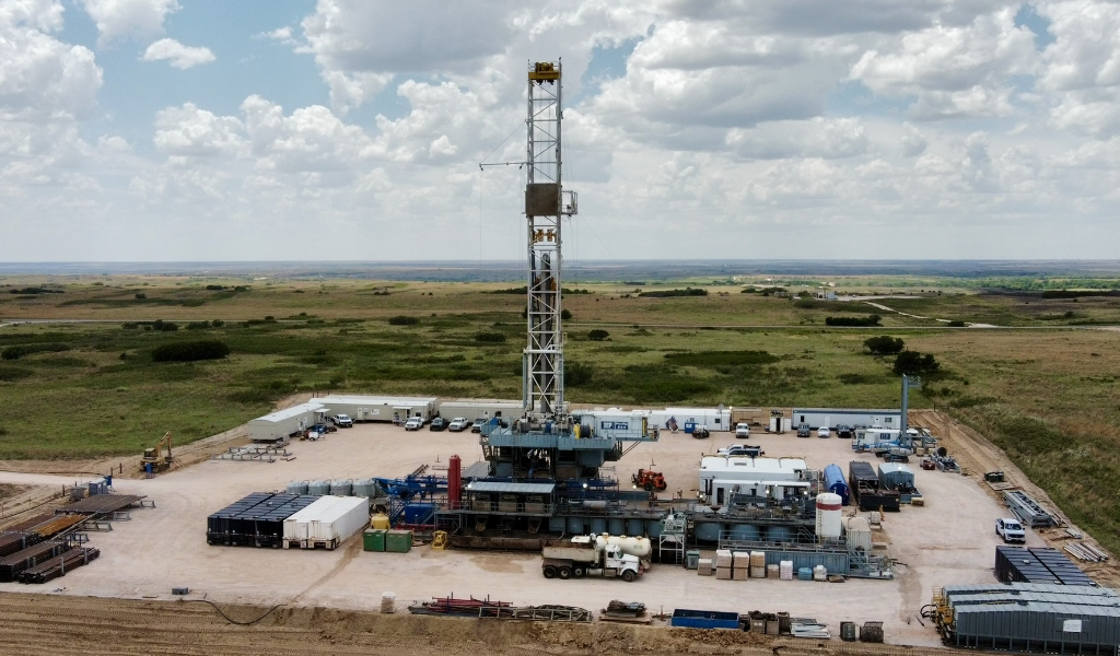 Aerial view of an oil drilling rig on a flat landscape, surrounded by equipment, vehicles, and storage containers under a partly cloudy sky.