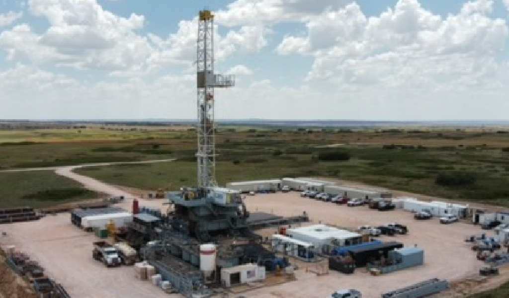 An oil drilling rig on a flat landscape, surrounded by equipment, vehicles, and storage containers under a partly cloudy sky.