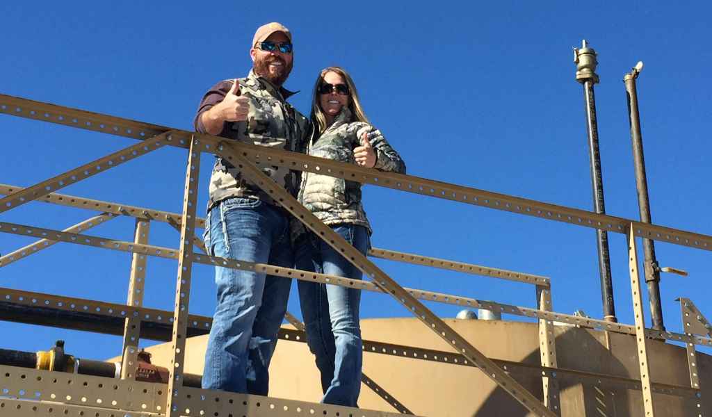 Two people in camouflage vests give thumbs up while standing on an industrial structure against a clear blue sky.